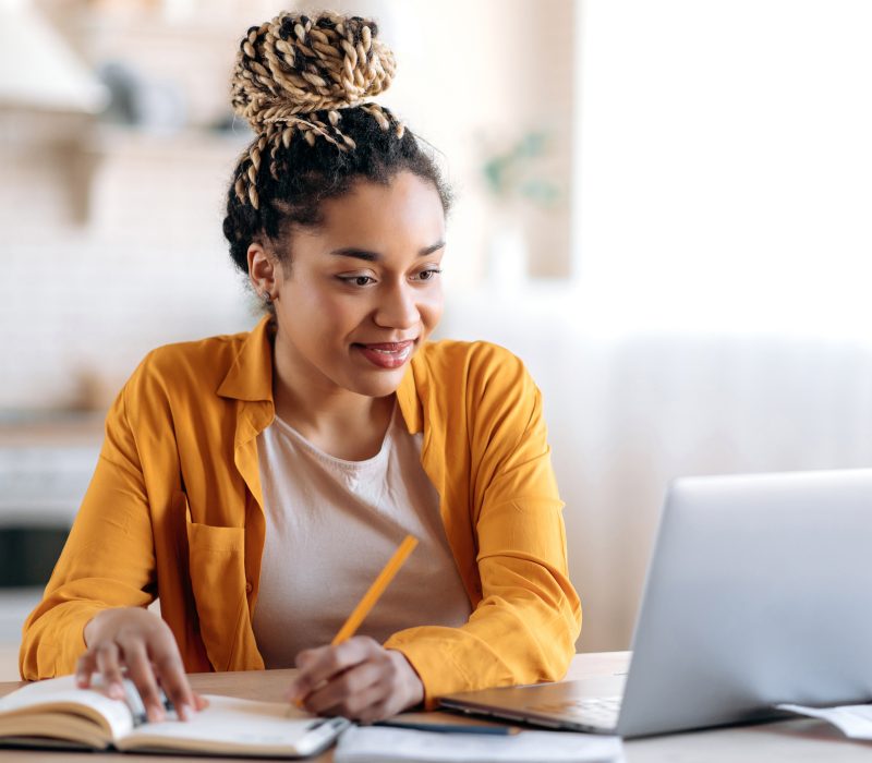 Focused cute stylish african american female student with afro dreadlocks, studying remotely from home, using a laptop, taking notes on notepad during online lesson, e-learning concept, smiling