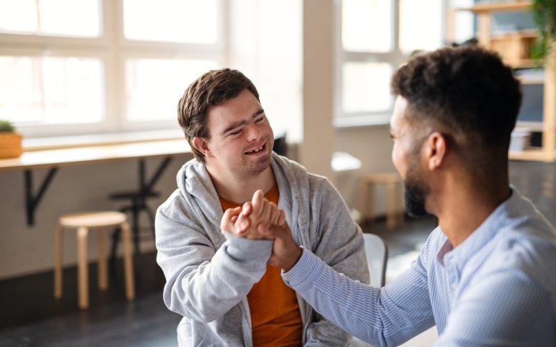 A young happy man with Down syndrome with his mentoring friend celebrating success indoors at school.