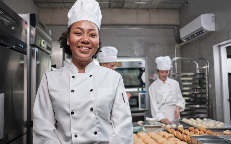 Portrait of young African American female chef in white cooking uniform looking at camera, cheerful smile with foods professional occupation, commercial pastry culinary jobs in a restaurant kitchen.