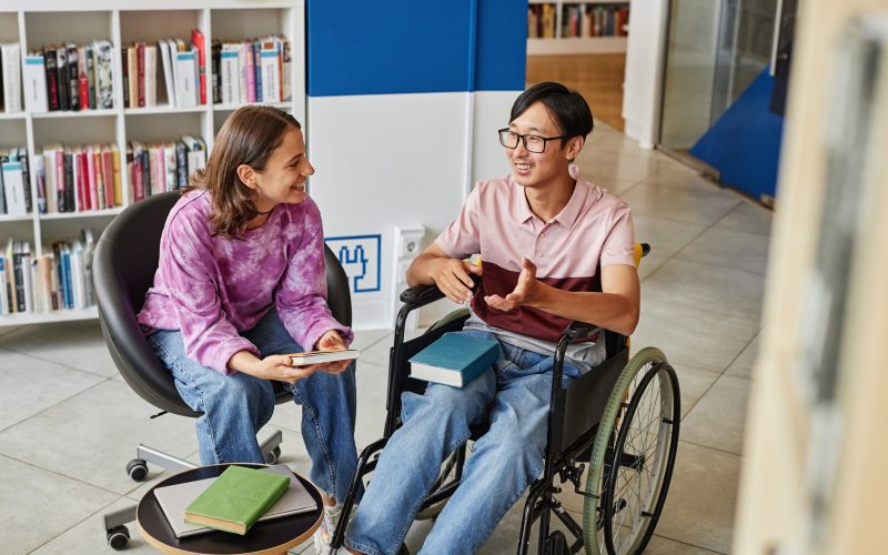 High angle portrait of Asian young man in wheelchair talking to friend while studying together in library