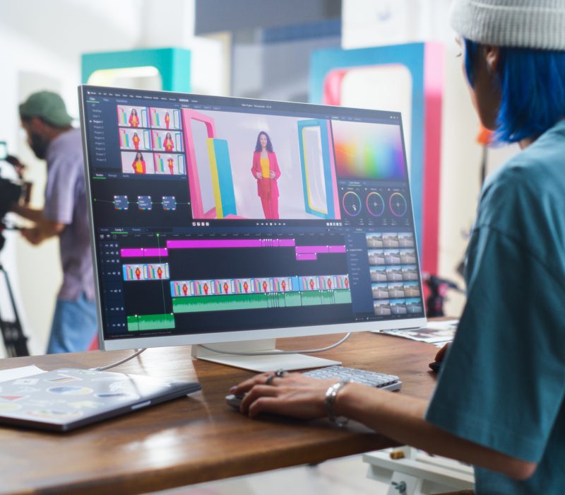 Young Caucasian Female Editor With Blue Hair Works On Video Editing Software, While A Hispanic Male Videographer Records A Black Female Host In A Vibrant Studio Setting, Enhancing Media Production.