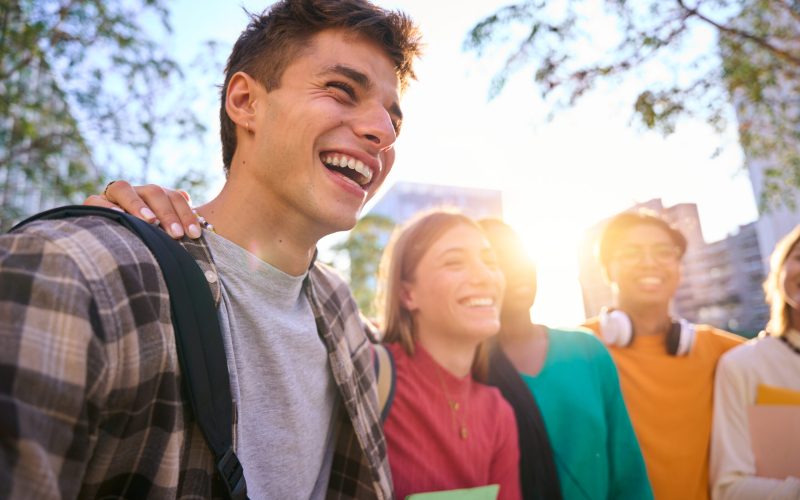 Happy caucasian guy with multiracial classmates university students posing together taking a photo. Generation z friends smiling and having fun holding backpacks outside the faculty buildings